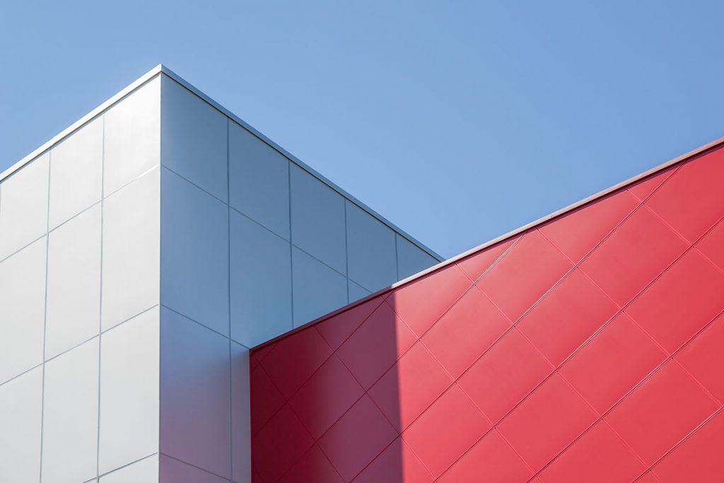 Top of a school building with a silver metallic and red meta l cladding. There is shadow in the building and a blue sky.